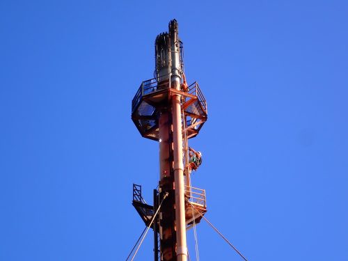 A worker in an orange high-visibility suit climbs a flare support structure against a clear blue sky. The structure is painted orange, matching the worker's attire, and is equipped with safety railings and platforms. The worker is secured with safety harnesses and ropes, indicating adherence to strict safety protocols. The photograph is taken from ground level, looking up and emphasising the structure's height and scale within the expansive sky's expansive backdrop.