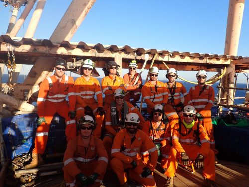 A group of industrial workers on an offshore platform pose for a photo. They wear bright orange coveralls, safety helmets, and protective glasses. Most are standing, and some are kneeling, with smiles indicating a moment of camaraderie or completing a job well done. The background features the metal structures of the platform, painted with visible rust, and the ocean extending to the horizon, suggesting a remote and harsh working environment. The clear sky and natural daylight enhance the visibility of the team's features and safety gear.