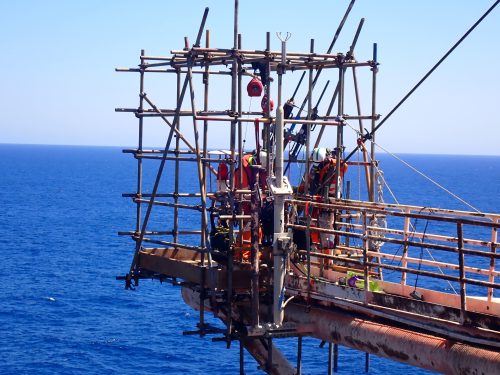 Industrial workers work in construction or maintenance on an offshore platform above the sea. They are working on a metal scaffold structure, secured with safety harnesses and wearing helmets for protection. The ocean provides a deep blue background, and the weather appears clear and sunny. The scene reflects the complexity and risk involved in offshore operations, where safety and precision are crucial due to the challenging environment.
