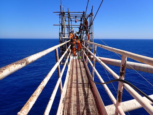 Workers in orange safety gear are on a narrow metal walkway above the open sea, with a clear blue sky. The walkway, showing signs of rust and wear, is supported by diagonal beams and leads to a scaffolding structure, indicating ongoing maintenance work. The workers are equipped with safety harnesses and helmets, adhering to safety regulations while working in this challenging and exposed offshore environment. The vast ocean stretches to the horizon, emphasising their work's isolated and expansive setting.