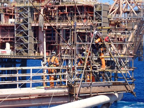 Workers clad in safety gear, including orange coveralls and white helmets, engage in offshore oil rig maintenance activities. They are surrounded by a complex lattice of metal beams, pipes, and safety railings, indicative of the rig's intricate infrastructure. The sea provides a vivid blue backdrop, highlighting the offshore environment where such industrial operations occur. The focus and coordination of the team suggest a high level of skill and attention to safety in a challenging work setting.