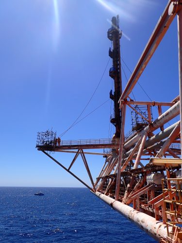 Workers in orange safety gear are gathered on a suspended metal platform against a clear blue sky and ocean backdrop. They appear to be engaged in a construction or maintenance task on the framework of an offshore structure. The platform is supported by rust-streaked beams that crisscross below it, contributing to a sense of height and open space around the workers. The calm sea extends to the horizon, emphasising the isolated and expansive environment where these offshore activities occur.