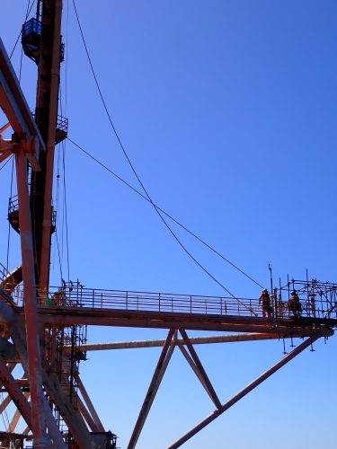 Workers in orange safety gear are gathered on a suspended metal platform against a clear blue sky and ocean backdrop. They appear to be engaged in a construction or maintenance task on the framework of an offshore structure. The platform is supported by rust-streaked beams that crisscross below it, contributing to a sense of height and open space around the workers. The calm sea extends to the horizon, emphasising the isolated and expansive environment where these offshore activities occur.