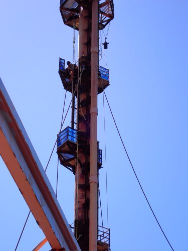 A towering metal structure with platforms, ladders, and safety railings rises against a clear blue sky. The structure, exhibiting a weathered rust patina, is equipped with pulleys and cables, indicative of its use for lifting or adjusting heavy equipment. The perspective is from the ground, looking up the structure's full height, emphasising its significant elevation and the work's verticality.