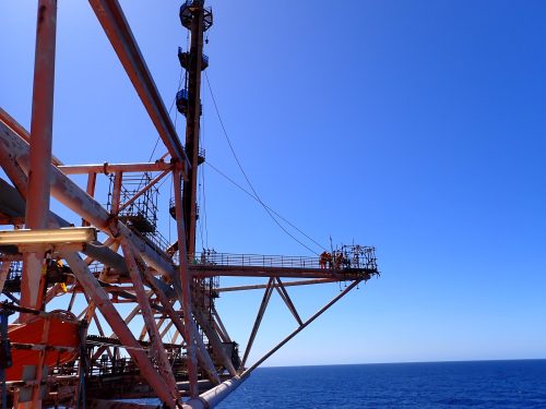 Industrial workers are perched on a scaffolding structure extending from a large, complex rig against the vast expanse of the ocean and under a clear blue sky. The structure is a lattice of rusted metal, and it includes safety features such as railings and stable platforms. Cables run alongside the scaffolding for additional support. This image captures the scale of offshore operations and the challenging environments workers face, emphasising the verticality and precariousness of their daily tasks.