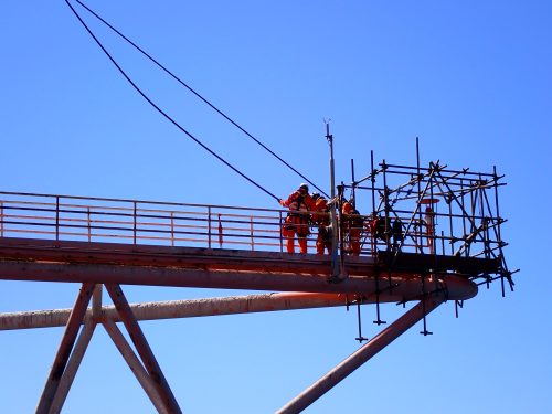 A group of workers in high-visibility orange suits and white helmets are on a metal scaffolding structure high above the ground. The scaffold is supported by a large rusted metal beam that angles upwards, with cables attached for added stability. They are working against an almost cloudless sky, which highlights the height at which they are working. This industrial scene occurs on an offshore oil rig structure requiring elevated construction or maintenance work.