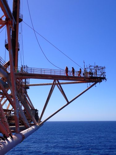 Workers in orange safety gear are gathered on a suspended metal platform against a clear blue sky and ocean backdrop. They appear to be engaged in a construction or maintenance task on the framework of an offshore structure. The platform is supported by rust-streaked beams that crisscross below it, contributing to a sense of height and open space around the workers. The calm sea extends to the horizon, emphasising the isolated and expansive environment where these offshore activities occur.