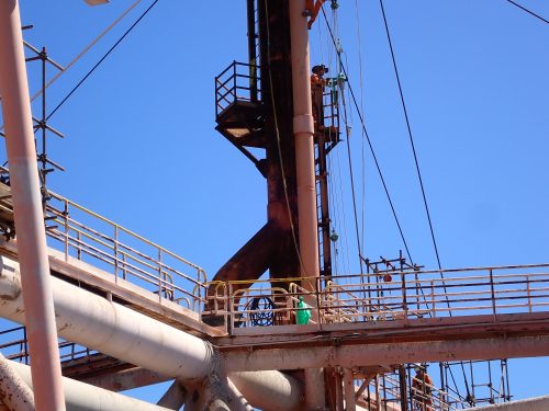 An industrial worker, secured with safety gear, climbs a vertical ladder on a large rust-coloured metal structure, part of an industrial or offshore facility. Metal pipes, railings, and cables parallel to the ladder are visible. The sky is clear, indicating good weather conditions for outdoor work. The perspective is from below, looking up towards the worker, highlighting the height and scale of the structure.