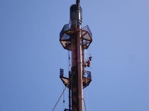 A worker in an orange high-visibility suit climbs a flare support structure against a clear blue sky. The structure is painted orange, matching the worker's attire, and is equipped with safety railings and platforms. The worker is secured with safety harnesses and ropes, indicating adherence to strict safety protocols. The photograph is taken from ground level, looking up and emphasising the structure's height and scale within the expansive sky's expansive backdrop.