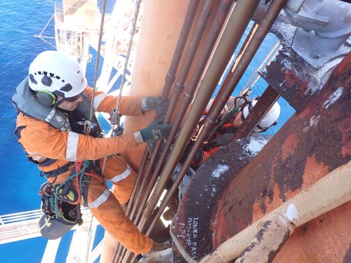 An industrial worker in orange safety gear, including a helmet with a mounted light and hearing protection, works on a vertical structure above the ocean. The worker is harnessed and using a descent control device attached to a rope, indicating rope access work. They appear to be inspecting or repairing a series of pipes on the side of the structure, which is streaked with rust, showing the harsh maritime environment. Below, the blue sea stretches out, emphasising the height at which the worker is operating.