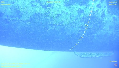 Underwater image captured by a remote-operated vehicle showing a section of the "Ichthys Venturer" hull with distinct marine growth and measurement markers. Text annotations include "INPEX", "Ichthys Venturer", and technical inspection details with date, time, heading, and depth information. The blue tint of the water creates a gradient effect, with the bottom of the hull and marine life more visible in the foreground.