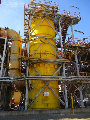 a large yellow cylindrical piece of refinery equipment covered in pipes and scaffolding.loading=