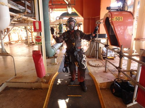 A vertech technician poses for a photo in a black protective suit and full face mask.