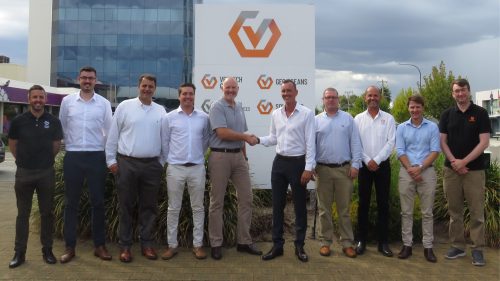 A group of ten men in business attire stood in front of a corporate building, with two in the centre shaking hands. They're posed before a sign with the Vertech Group Logo with Group companies below, and the setting is daytime with partly cloudy skies and some greenery around them.