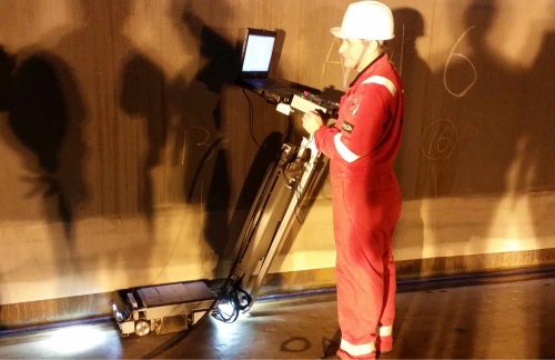 A technician in red coveralls with reflective strips and a white hard hat operates a portable, wheeled ultrasonic testing device connected to a laptop known as an MEC Scanner. They are inspecting a large metallic surface, likely the wall of a tank or vessel, which has handwritten numbers indicating inspection areas. The environment is dimly lit, with shadows cast by the equipment and the worker, who appears focused on the inspection process, showcasing an industrial non-destructive testing (NDT) operation in progress.