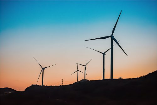 A silhouette of wind turbines on a hill against a twilight sky. The gradient of the sky fades from a deep blue at the top to a warm orange near the horizon. The turbines are prominently outlined and spaced across the rolling landscape, capturing a moment of tranquillity and the sustainable harnessing of wind energy during dusk.