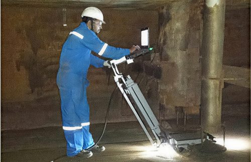 An operator in blue coveralls and a white safety helmet uses a sophisticated digital display inspection device for ultrasonic testing known as a MEC Scanner. The technician is focused on the device screen while the scanner is operating. The setting is an industrial environment with visible metal beams and structures. The scene is illuminated by artificial light sources that glow on the floor, emphasising the inspection task in a dimly lit area.