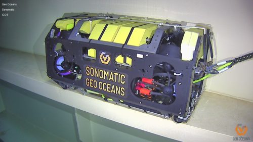 A high-tech, industrial piece of equipment, the i-COT is a remotely operated ballast tank inspection device with the words "SONOMATIC GEO OCEANS" printed on the side. It has a robust, dark frame with yellow and orange components, wires, and cylindrical parts, indicative of sophisticated marine technology. The device rests on a shelf indoors, and a cable is connected to it, extending off the right side of the frame.