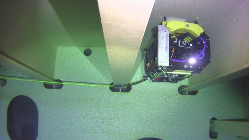 The remotely operated ballast tank inspection device, i-COT, is attached to a green cable and navigates near a large submerged structure with various openings. The equipment features yellow accents and is equipped with lights, suggesting active operation. It is in the midst of inspection or data collection. The surrounding environment is dimly lit, which is characteristic of underwater conditions.