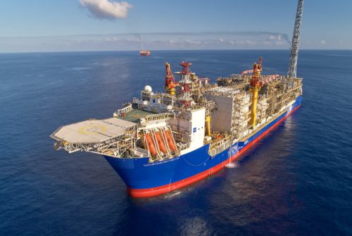 An aerial view showcases a colossal floating production storage and offloading (FPSO) vessel anchored in the open ocean. Its hull is painted in vibrant blue with a red stripe, while the deck is loaded with intricate, multicoloured industrial machinery for processing oil and gas. The vessel features a sprawling helipad, towering cranes, and a massive processing plant. A long boom extends from the vessel's aft, hinting at its offshore drilling operations. The sea around the boat reflects the sunlight and a distant support ship can be seen on the horizon under a clear sky with a few scattered clouds.