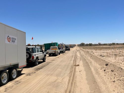 A convoy of vehicles, including a four-wheel drive and trucks with safety flags, is parked along a dirt road in a sparse, arid landscape. The leading car tows a large grey trailer emblazoned with the Vertech Group logo. The road stretches into the distance under a clear blue sky, flanked on one side by a low fence and on the other by open, flat terrain with sparse vegetation. The scene suggests an industrial or maintenance activity in a remote, desert-like environment.