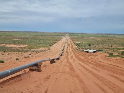 An expansive view of a flat, arid landscape with a long dirt road stretching straight into the distance. Parallel to the road, a large metal pipeline is supported at intervals by wooden beams. A white pickup truck is parked beside the pipeline, dwarfed by the vastness of the terrain. The open sky is partially cloudy with hints of blue, and the horizon line is unbroken, save for the faint suggestion of distant vegetation. The scene indicates infrastructure development in a remote, sparsely vegetated desert area.