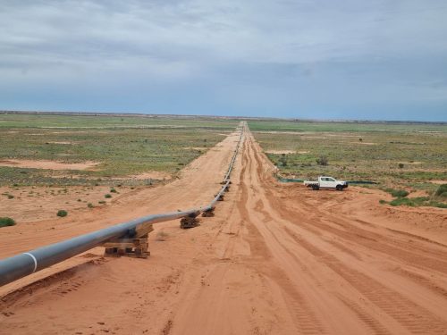 A vast, flat expanse of arid land stretches into the distance, marked by a straight, unsealed road cutting through the landscape. Parallel to the road runs a large metal pipeline, supported intermittently by wooden blocks. A white utility vehicle is parked beside the pipeline, its presence minuscule in the vastness of the terrain. The environment is characterised by red soil, sparse green shrubbery, and a wide horizon under a partly cloudy sky, evoking a sense of remote industrial activity in a desert-like setting. A vast, flat expanse of arid land stretches into the distance, marked by a straight, unsealed road cutting through the landscape. Parallel to the road runs a large metal pipeline, supported intermittently by wooden blocks. A white utility vehicle is parked beside the pipeline, its presence minuscule in the vastness of the terrain. The environment is characterised by red soil, sparse green shrubbery, and a wide horizon under a partly cloudy sky, evoking a sense of remote industrial activity in a desert-like setting.