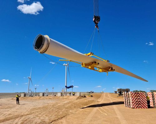 A large wind turbine blade is hoisted by a crane against a vivid blue sky with a single cloud. In the background, other turbines and construction equipment are visible on the wind farm. A worker wearing high-visibility clothing oversees the operation from the ground, ensuring safety and precision in the installation process. The photo captures the scale and scope of renewable energy infrastructure projects in a rural setting.