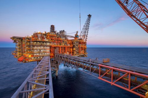 An offshore oil platform is illuminated against the twilight sky. The sprawling structure is a complex of pipes, towers, and equipment extending into the sea. A prominent flare structure rises on the right, mirrored by the bridge connecting the platform to the smaller structures on the left. The sky transitions from warm sunset hues to the deep blue of the approaching night while the calm sea stretches to the horizon, reflecting the last light of day. The image captures the industrial might set against the natural beauty of the ocean at dusk.