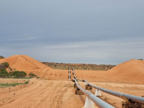 A landscape of a mining site with a series of pipes running parallel to a dirt road, extending toward the horizon. Large mounds of reddish-brown earth pile up on either side of the pathway, indicating recent excavation or construction. The sky is overcast with greyish clouds, suggesting a possible storm approaching, while sparse vegetation dots the otherwise barren and sandy terrain.