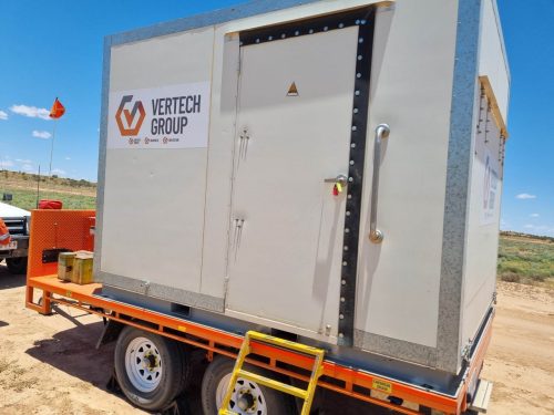 A large mobile container unit is mounted on a dual-axle trailer parked on sandy terrain under a clear sky. The side of the container displays the Vertech Group logo. An orange safety flag on a pole and a diamond-shaped chemical hazard sign are visible on the left. The trailer is equipped with off-road tyres suitable for the terrain, and a sturdy metal step provides access to the container's door. The shadows on the ground indicate bright overhead sunlight, which is common in outback environments.