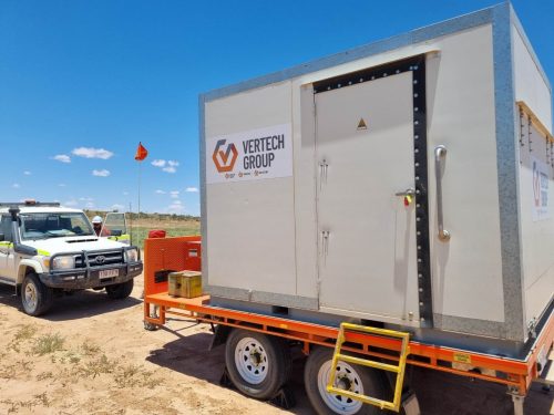The image shows a sturdy, white container with the "Vertech Group" logo mounted on an orange and silver trailer, hinting at industrial or technical use. It is parked on sandy ground, indicative of a remote or rural location, with a white Toyota ute in the background bearing safety flags, commonly used in industrial or construction sites to enhance vehicle visibility. The clear blue sky and bright sunlight suggest a dry, sunny day. The container's solid build and the presence of safety equipment imply adherence to occupational health and safety standards for field operations.
