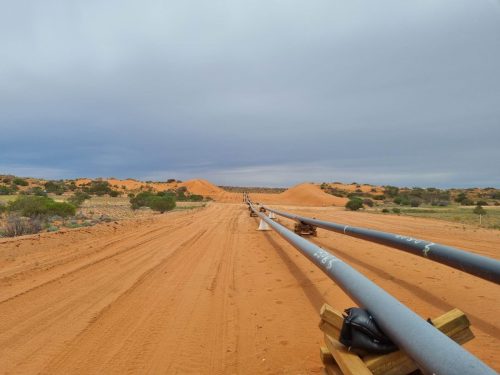 A view of a sandy, unpaved road cutting through a semi-arid landscape dotted with sparse vegetation and red dunes under an overcast sky. The road is flanked on one side by a long stretch of pipe supported by wooden beams, suggesting ongoing construction or maintenance work possibly related to energy or resource extraction. The pipes extend into the horizon, emphasising the scale of the project within the remote and expansive environment.