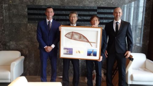 Four individuals in business attire stand in a modern office, holding a large framed artwork between them. The artwork has an indigenous Australian model of a fish, with smaller images beneath it, possibly related to the subject or an event. The group poses for a commemorative photograph, two men on the sides and two in the middle holding the frame. They all exhibit a professional demeanour, and the environment suggests a formal occasion, possibly a presentation or an award ceremony.